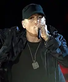 A man in a black cap, t-shirt and jacket, and dog tag performing with a microphone in his hand