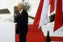 A side view of Jim Robson speaking to a crowd and holding a microphone in his right hand