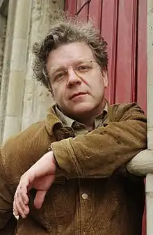 Middle-aged white male wearing glasses and brown button-up jacket, leaning on a wall