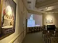 The St. George Visitors Center theatre, playing a movie on the history of the temple. Chairs are shown in the foreground, to the side are two paintings of the life of Jesus Christ.